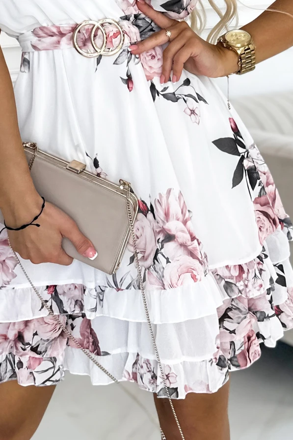 Elegantes weißes Kleid mit floralen Mustern und einem doppelt geschnallten Gürtel, das einen femininen und stilvollen Look bietet. Die Kombination aus zarten Rüschen und einer kleinen, beigen Handtasche verleiht dem Outfit eine romantische Note.
