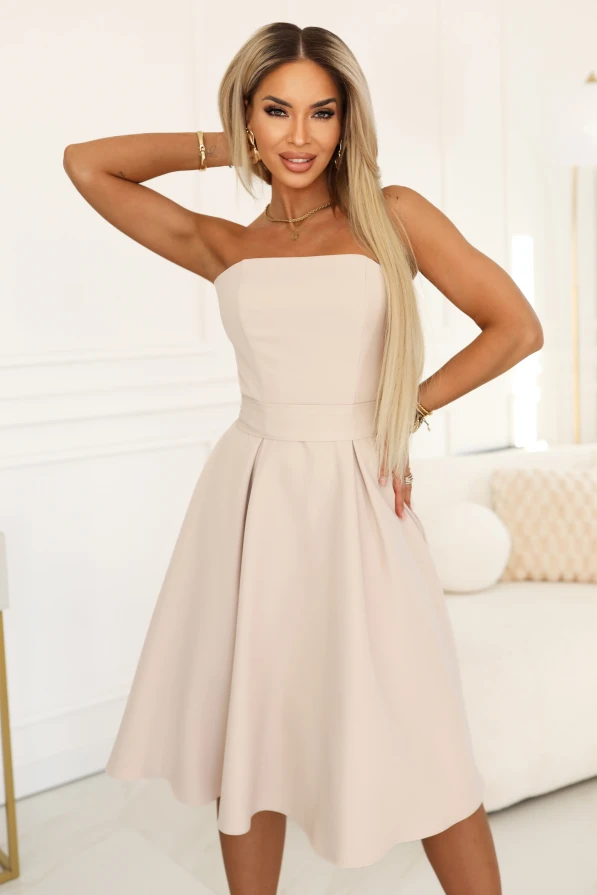 Das Model präsentiert ein elegantes hellbeiges trägerloses Midiekleid mit ausgestelltem Rock und betonter Taille, ideal für Hochzeiten, Kommunionen und Empfänge.