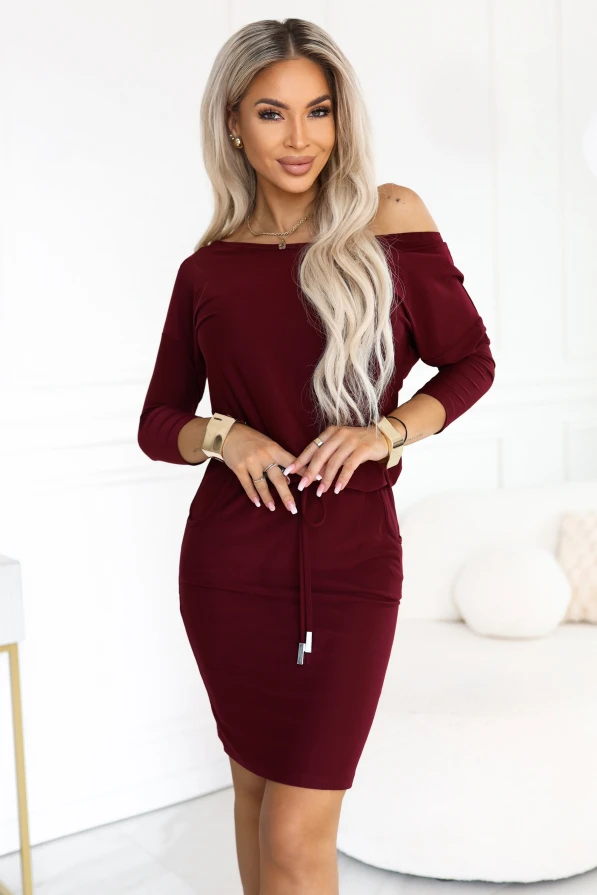 Model in einem bordeauxfarbenen sportlichen Numoco Kleid knapp über dem Knie mit Bootausschnitt, der eine Schulter freilässt, 3/4-Ärmeln, seitlichen Taschen und Bindung in der Taille.