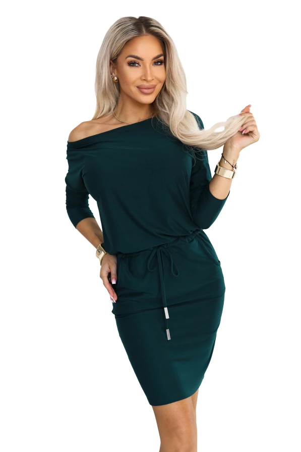 Model in einem sportlichen Kleid in Flaschengrün mit einem Ausschnitt, der eine Schulter freilässt, 3/4-Ärmeln, seitlichen Taschen und Bindung in der Taille. Schnitt mit lockerem Oberteil und figurbetontem Unterteil knapp über dem Knie.