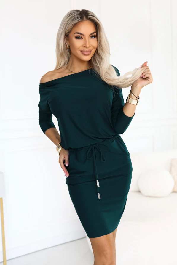 Model in einem sportlichen Kleid in Flaschengrün mit einem Ausschnitt, der eine Schulter freilässt, 3/4-Ärmeln, seitlichen Taschen und Bindung in der Taille. Schnitt mit lockerem Oberteil und figurbetontem Unterteil knapp über dem Knie.