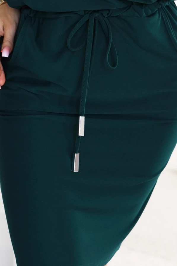 Model in einem sportlichen Kleid in Flaschengrün mit einem Ausschnitt, der eine Schulter freilässt, 3/4-Ärmeln, seitlichen Taschen und Bindung in der Taille. Schnitt mit lockerem Oberteil und figurbetontem Unterteil knapp über dem Knie.