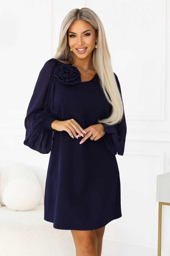 Model im marineblauen Kleid NELIA von Numoco im Trapezschnitt knapp über dem Knie, mit dekorativer Rose am Ausschnitt und langen, transparenten Ärmeln, die mit breiten Manschetten mit Knopf abschließen.