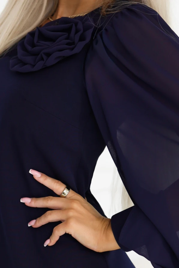 Model im marineblauen Kleid NELIA von Numoco im Trapezschnitt knapp über dem Knie, mit dekorativer Rose am Ausschnitt und langen, transparenten Ärmeln, die mit breiten Manschetten mit Knopf abschließen.