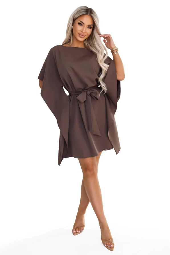 Model im Kleid SOFIA von Numoco in einem Mokkabraun-Ton. Das Kleid reicht knapp über das Knie, hat einen Bootausschnitt, weite Schmetterlingsärmel und einen in der Taille zur Schleife gebundenen Gürtel.