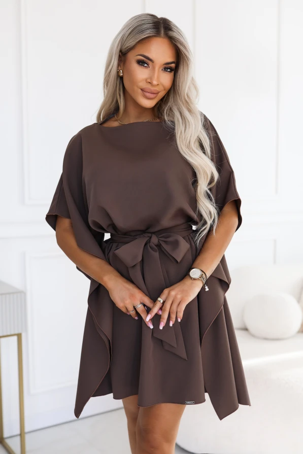 Model im Kleid SOFIA von Numoco in einem Mokkabraun-Ton. Das Kleid reicht knapp über das Knie, hat einen Bootausschnitt, weite Schmetterlingsärmel und einen in der Taille zur Schleife gebundenen Gürtel.
