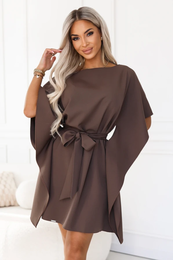 Model im Kleid SOFIA von Numoco in einem Mokkabraun-Ton. Das Kleid reicht knapp über das Knie, hat einen Bootausschnitt, weite Schmetterlingsärmel und einen in der Taille zur Schleife gebundenen Gürtel.