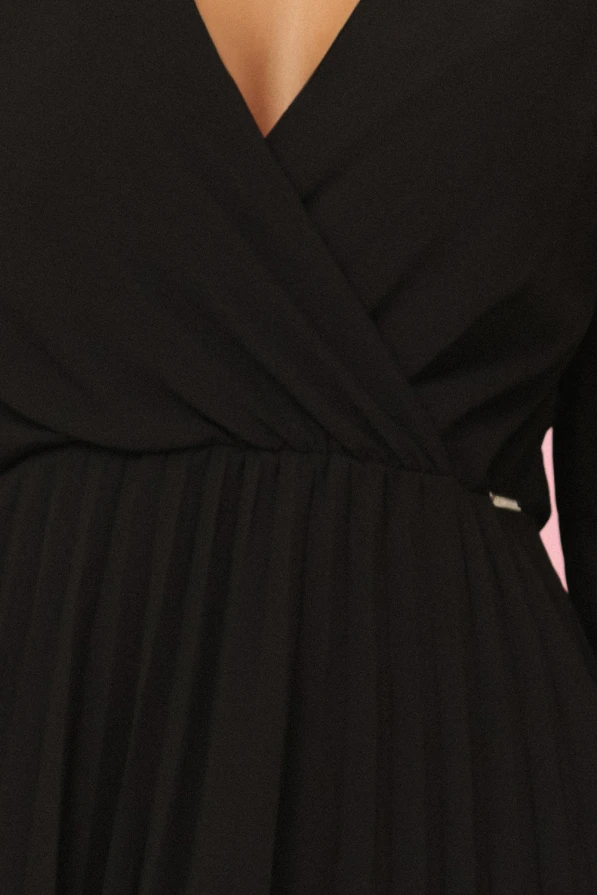 Modelka prezentuje elegancką czarną szyfonową sukienkę marki Numoco z długim rękawem, kopertowym dekoltem i plisowanym dołem – idealną na wieczór, wesele lub specjalne okazje. Produkt polski, sfotografowany na różowym tle.