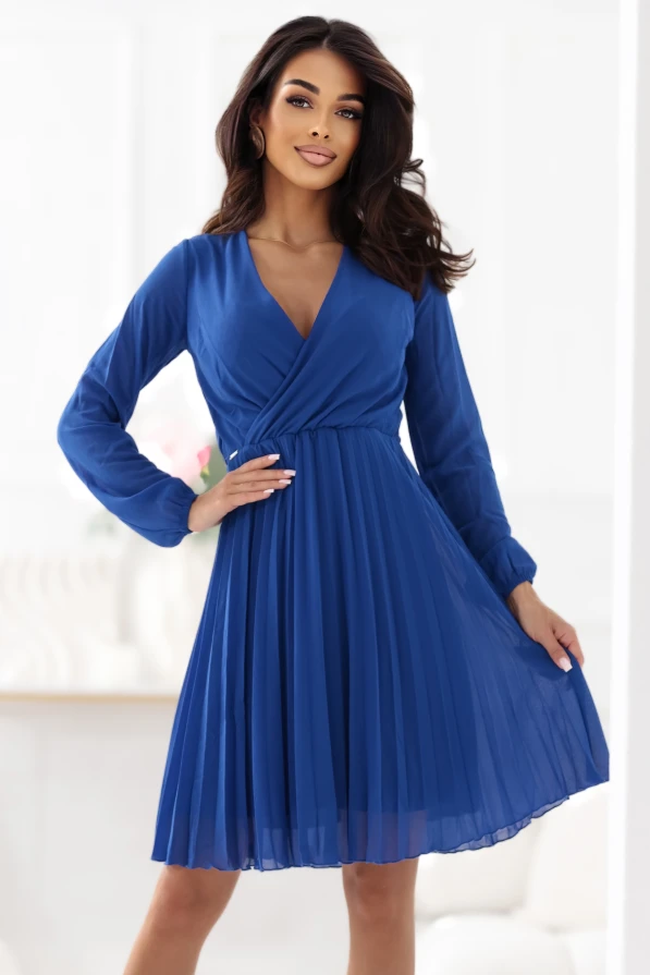 Modelka prezentuje elegancką chabrową sukienkę z kopertowym dekoltem i plisowanym dołem. Stylizacja marki Numoco została sfotografowana w jasnym, nowoczesnym wnętrzu, co podkreśla lekkość i szyk kreacji.