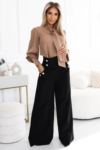 Eine elegante Frau trägt eine stilvolle Bluse in Beige mit einer Schleife und weiten, schwarzen Hosen mit auffälligen Knöpfen. Das Outfit strahlt Raffinesse und modernen Chic aus, perfekt für formelle Anlässe.}