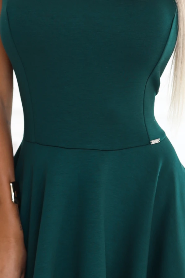 Modelka prezentuje elegancką sukienkę w kolorze butelkowej zieleni z rozkloszowanym dołem i dłuższym tyłem, stojąc w jasnym, stylowym wnętrzu. Kreacja podkreśla kobiecą sylwetkę i doskonale nadaje się na wyjątkowe okazje.