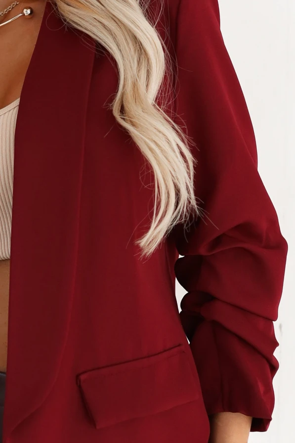 Elegancka stylizacja damska – modelka w bordowej marynarce, beżowym topie i skórzanej mini spódnicy, dopełniona kapeluszem i długimi kozakami; modna jesienna stylizacja w jasnym, minimalistycznym wnętrzu.