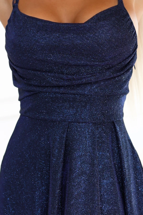 Modelka prezentuje elegancką granatową sukienkę maxi z połyskiem, dopasowaną górą i efektownym rozcięciem na nodze. Kreacja z wiązaniem na plecach podkreśla sylwetkę i sprawdza się jako suknia wieczorowa na wyjątkowe okazje.