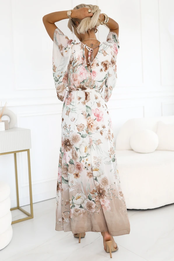 Modelka prezentuje zwiewną sukienkę maxi z kwiatowym printem w odcieniach beżu i różu, z kopertowym dekoltem, luźnymi rękawami i paskiem w talii. Idealna propozycja na wesele, przyjęcie lub letni spacer – lekka, kobieca i efektowna.