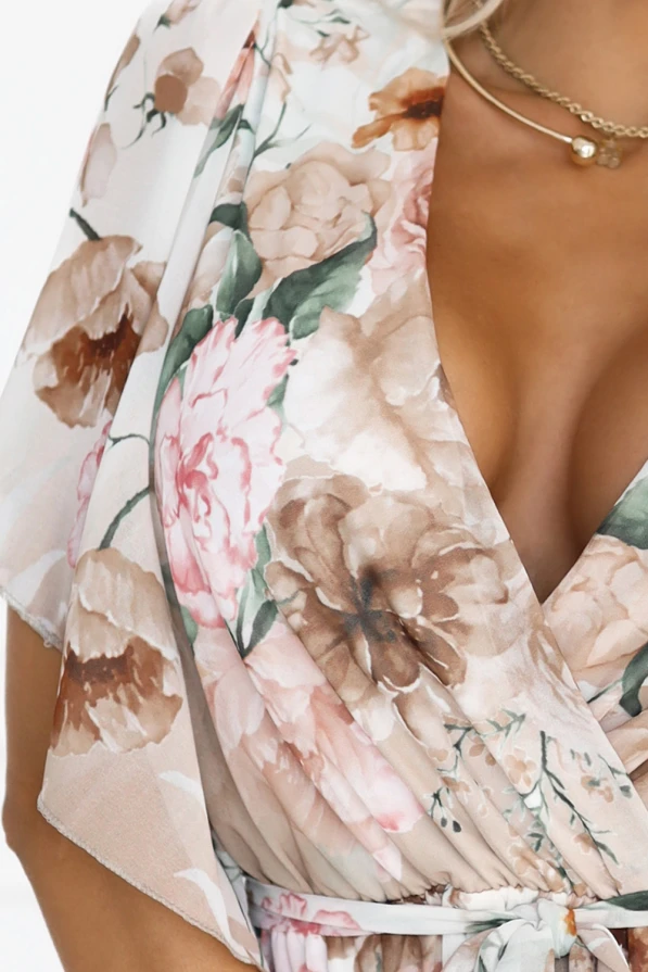 Modelka prezentuje zwiewną sukienkę maxi z kwiatowym printem w odcieniach beżu i różu, z kopertowym dekoltem, luźnymi rękawami i paskiem w talii. Idealna propozycja na wesele, przyjęcie lub letni spacer – lekka, kobieca i efektowna.