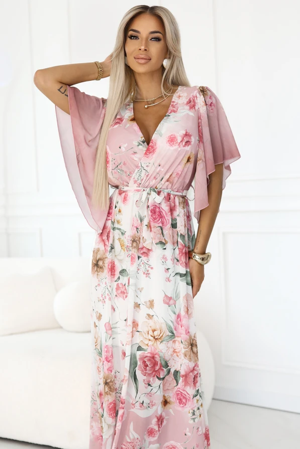 Modelka prezentuje zwiewną sukienkę maxi w romantyczny, kwiatowy wzór w odcieniach różu. Jej subtelna poza podkreśla lekkość tkaniny oraz kobiecy fason z wiązaniem w pasie i luźnymi rękawkami.