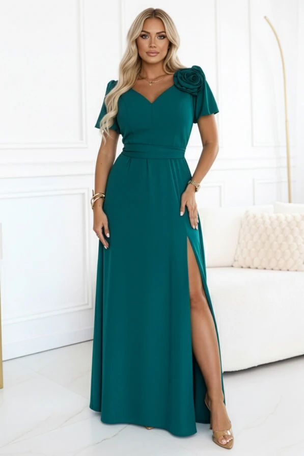Butelkowozielona długa sukienka z ozdobną różą i wysokim rozcięciem, zaprezentowana przez modelkę w jasnym, eleganckim wnętrzu. Plus size.